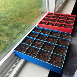 以下が含まれることがあります： 赤と青の2つのカラフルな種子トレイ。各トレイには16個の正方形のコンパートメントがあり、暗い土で満たされています。 トレイは窓枠に置かれ、植え付けの準備ができています。 背景には緑の芝生の景色が広がっています。