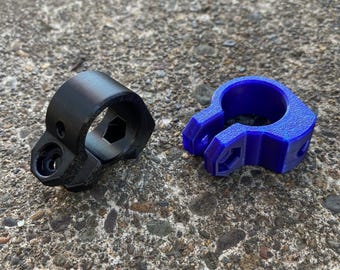 May include: Two 3D-printed tools. One is black, with a hexagonal opening and a small circular hole. The other is blue, with a similar hexagonal opening and a claw-like design. Both are on a textured gray surface.