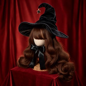 May include: A black velvet witch hat featuring a pointed, slightly slumped crown and a wide, curved brim. A small gold-toned star charm with a black centre hangs from the tip of the hat. The hat is displayed on a mannequin head with long, wavy brown hair and a black satin ribbon tied in a bow at the neck.