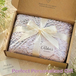 May include: A gift box with a white ribbon tied in a bow. The box contains a wrapped item with a patterned design. The box is open, revealing the contents. The text on the box reads "Thank you for your support. It means the world to us."