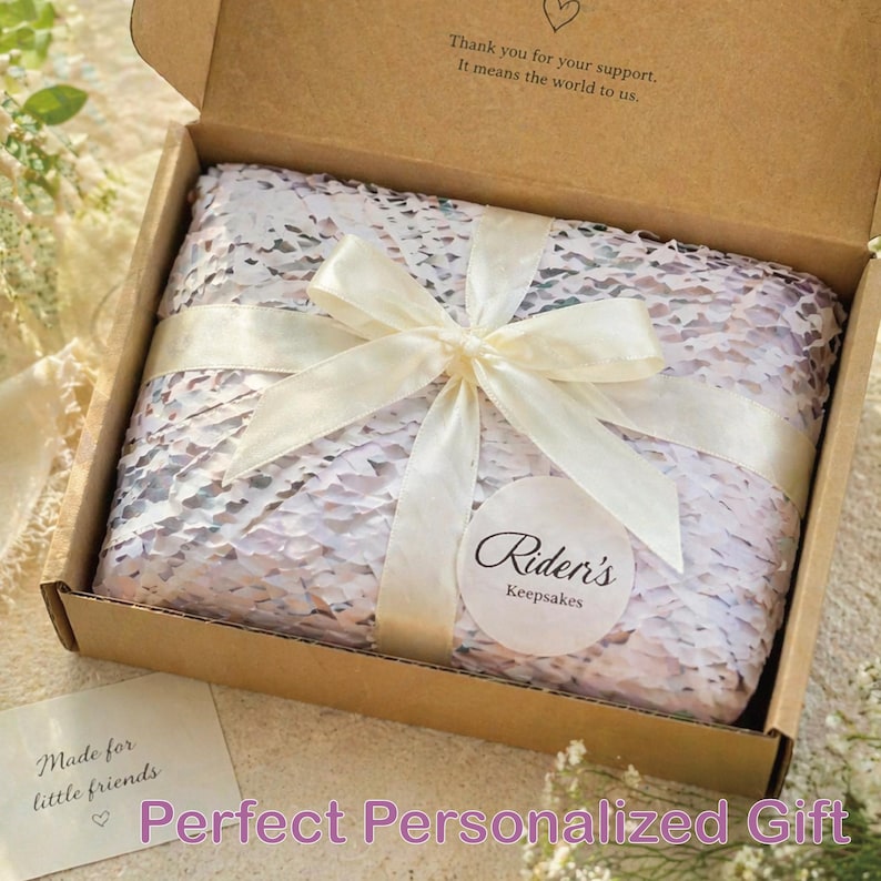 May include: A gift box with a white ribbon tied in a bow. The box contains a wrapped item with a patterned design. The box is open, revealing the contents. The text on the box reads "Thank you for your support. It means the world to us."