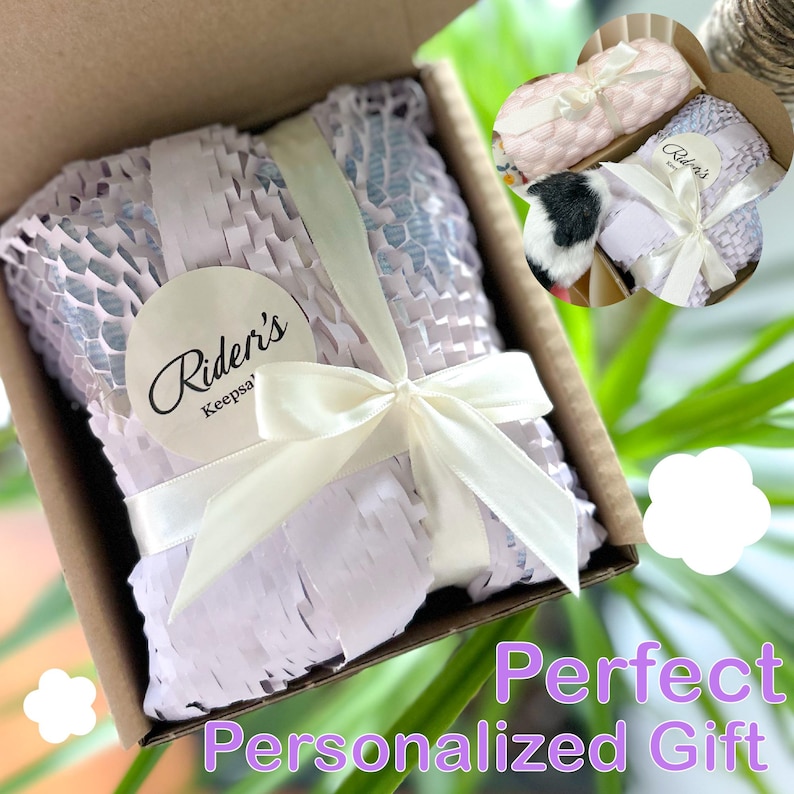 May include: A gift box with a cream-colored ribbon and bow. The box contains a wrapped item, protected by lavender-colored honeycomb packing material. A round sticker with the text "Rider's Keepsa'" is on the wrapping. The words "Perfect Personalized Gift" are at the bottom.