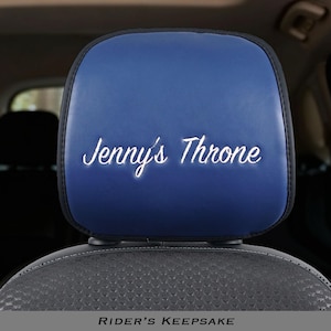 May include: A blue car headrest cover with black trim featuring the white embroidered text Jenny's Throne centered on the front surface. The cover is fitted over a vehicle headrest, displaying a smooth finish and clear, legible script lettering.