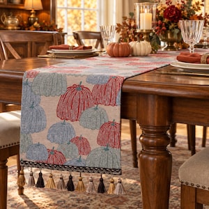 May include: A rectangular table runner featuring a woven pattern of red and blue pumpkins on a beige background. The runner is finished with a dark brown decorative trim and a row of multi-colored fabric tassels in shades of brown and tan.