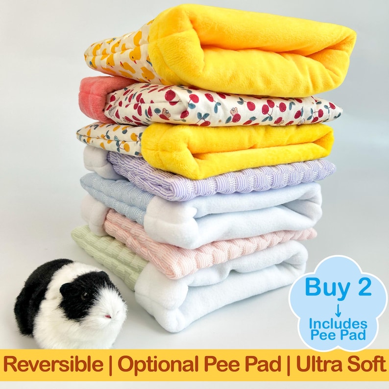 May include: A stack of colorful, reversible pet bed pads in various patterns and colors, including yellow, pink, and blue. The pads are stacked with a guinea pig in the foreground. Text on the image reads "Reversible | Optional Pee Pad | Ultra Soft".