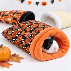 Könnte beinhalten: Ein schwarzer und orangefarbener Stoffschlafsack mit Kürbismuster für kleine Haustiere mit weichem, orangefarbenem Innenfutter. Ein schwarz-weißes Meerschweinchen schaut aus der Öffnung hervor. Zwei weitere Stoffbeutel sind im Hintergrund zu sehen.