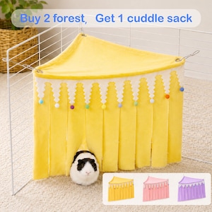 Può includere: Un nascondiglio angolare triangolare giallo in pile per piccoli animali con bordo bianco a zigzag e pon-pon colorati appesi. Il testo Buy 2 forest, Get 1 cuddle sack è visibile. Una cavia bianca e nera è posizionata davanti.