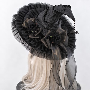 May include: A black witch hat featuring a crinkled texture, a wide ruffled lace brim, and two black fabric roses with matching bows. A dangling charm featuring small black beads and a bat silhouette hangs from the pointed tip of the hat.