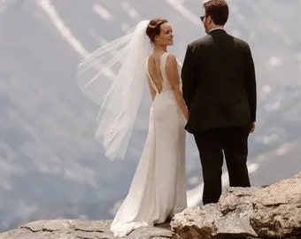 May include: A bride and groom stand on a rocky cliff overlooking a mountain range. The bride is wearing a white wedding dress with a veil and the groom is wearing a black suit. The couple is looking at each other and smiling.