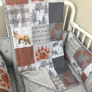 May include: A gray and brown baby crib bedding set with a fox, bear paw print, and trees. The bedding set includes a quilt, a pillow, and a bumper. The quilt has a patchwork design with different patterns and colors. The pillow and bumper are both gray with a white polka dot pattern. The text "WILD AND FREE" is repeated on the quilt.