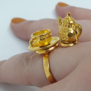 May include: A gold-tone ring with a miniature teapot and teacup attached. The teapot has a lid and a spout, and the teacup has a handle and a saucer.