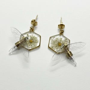 Puede incluir: Un par de pendientes dorados. Cada pendiente presenta un colgante hexagonal transparente de resina con una flor blanca seca y un diseño de insecto en miniatura. El insecto tiene alas transparentes y un cuerpo dorado.