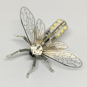 May include: A silver and white insect-shaped sculpture with intricate details. The wings are made of clear plastic with a glittery finish and feature circuit board patterns. The body is made of metal and includes a small LED light.