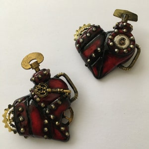 May include: Two red heart-shaped steampunk-style pendants with intricate metal gears, keys, and rivets. One pendant has the word "AMPER" on it.