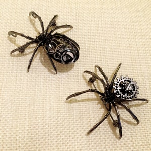 May include: Two black metal spider brooches with intricate gear details. One spider has a larger gear on its back, while the other has smaller gears throughout its body.