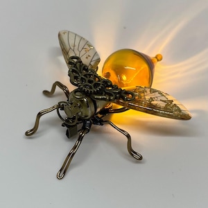 May include: A steampunk-style bee sculpture with a glowing amber glass body and delicate, patterned wings. The bee features intricate brass-colored metalwork, including gears and wire legs, creating a unique decorative piece.