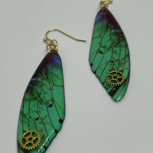 Op de afbeelding: Een paar unieke oorbellen met een groen, vleugelvormig ontwerp en gouden accenten. De vleugels hebben een kleurengradiënt met een vleugje paars en zwart. Elke oorbel heeft een kleine gouden tandwielbedel en een gouden haak.