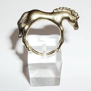 May include: A gold-colored horse ring. The horse is standing on its hind legs with its head turned to the side. The ring is on a clear acrylic stand.