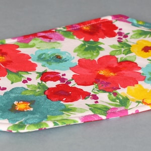 May include: A white fabric pot holder with a floral pattern featuring red, yellow, pink, blue, and green flowers. The pot holder is quilted and has a hexagonal shape.