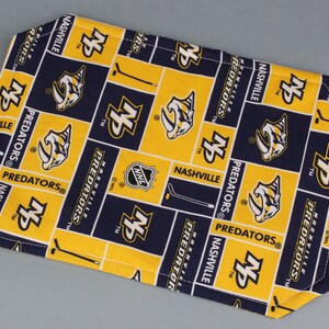 May include: A blue and yellow fabric placemat with the Nashville Predators logo repeated throughout. The logo features a stylized sabertooth cat with the letters "NS" and the words "Nashville Predators" repeated throughout.