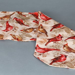 May include: A white table runner with a repeating pattern of red cardinals and brown pine branches. The runner has a scalloped edge.