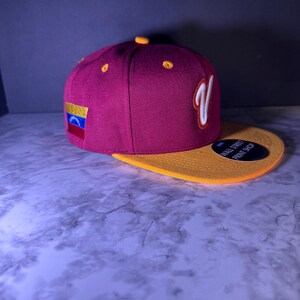 May include: A maroon and gold baseball cap with a flat brim. The cap features a white "V" logo and a small Venezuelan flag patch. The brim is gold, and the cap has the text "WALL STREET PRINT SHOP".