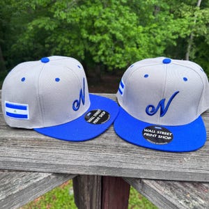 May include: Two gray baseball caps with blue brims are displayed. Each cap features a blue embroidered letter "N" and a small flag on the side. The text "WALL STREET PRINT SHOP" is printed on a sticker on the brim.