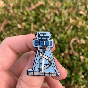 May include: A blue and white enamel pin depicting an airport control tower with a spiral staircase. The pin is a detailed representation of a control tower.