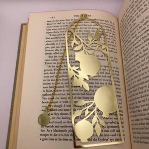 Op de afbeelding: Goudkleurige metalen bladwijzer met een granaatappelontwerp. De rechthoekige bladwijzer heeft een decoratieve uitsparing van een granaatappeltak en een bungelende bedel. Het ligt op de pagina's van een boek.