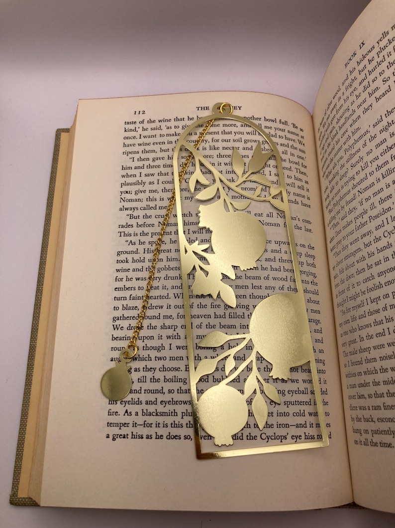 Op de afbeelding: Goudkleurige metalen bladwijzer met een granaatappelontwerp. De rechthoekige bladwijzer heeft een decoratieve uitsparing van een granaatappeltak en een bungelende bedel. Het ligt op de pagina's van een boek.