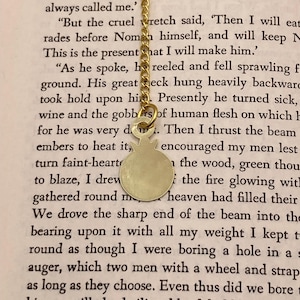 Op de afbeelding: Een goudkleurige hangerketting met een ronde bedel en een fijne ketting. De bedel heeft een eenvoudig, minimalistisch ontwerp. De ketting wordt getoond tegen een achtergrond van gedrukte tekst.