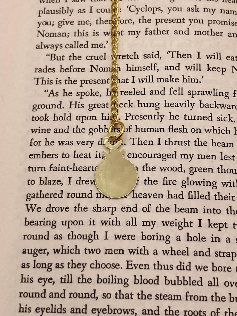 Op de afbeelding: Een goudkleurige hangerketting met een ronde bedel en een fijne ketting. De bedel heeft een eenvoudig, minimalistisch ontwerp. De ketting wordt getoond tegen een achtergrond van gedrukte tekst.