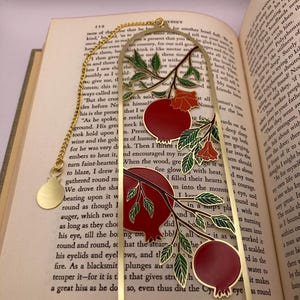 Op de afbeelding: Goudkleurige metalen bladwijzer met een granaatappelontwerp. Het heeft rode vruchten, groene bladeren en een gouden ketting. Rechthoekig met een gewelfde bovenkant, liggend op een open boek.