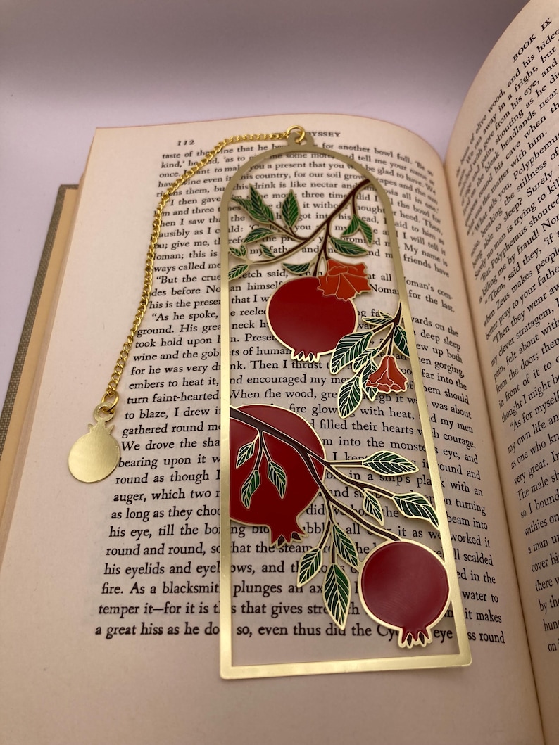 Op de afbeelding: Goudkleurige metalen bladwijzer met een granaatappelontwerp. Het heeft rode vruchten, groene bladeren en een gouden ketting. Rechthoekig met een gewelfde bovenkant, liggend op een open boek.