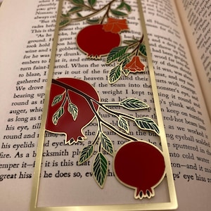 Op de afbeelding: Een goudkleurige metalen bladwijzer met een granaatappelontwerp. De bladwijzer heeft rode granaatappels, groene bladeren en oranje bloemen. De bladwijzer ligt op de pagina's van een boek.