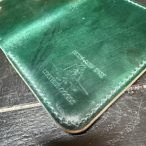 Op de afbeelding: Een groene leren portemonnee met witte stiksels rond de randen. De portemonnee heeft een logo en de tekst "LAND AND SON LEATHER GOODS". Het leer heeft een getextureerde uitstraling.