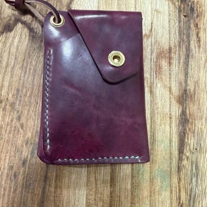 Op de afbeelding: Een bordeauxrode leren kaarthouder met een klepsluiting en messing ogen. De randen zijn gestikt met lichtgroen garen. Er is een leren lus bevestigd om te dragen. De kaarthouder ligt op een houten oppervlak.