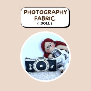 May include: A fabric doll with red braided hair, glasses, and a black and white dress, next to a black and white camera-shaped pillow. The text "PHOTOGRAPHY FABRIC (DOLL)" is displayed above the doll.