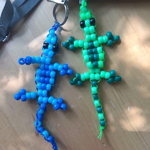 May include: Two beaded lizard keychains. One is blue with black eyes, the other is green with black eyes. Both have a beaded body, legs, and tail. They are attached to silver key rings.
