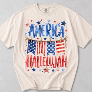 Peut inclure: Un t-shirt à manches courtes de couleur crème avec un motif aquarelle comprenant le texte bleu "AMERICA" au-dessus d'une guirlande de quatre fanions aux couleurs du drapeau américain, et le texte rouge "HALLELUJAH" avec des étoiles.