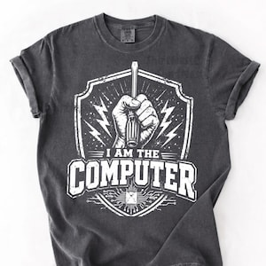 Peut inclure: T-shirt gris foncé avec un motif blanc. Le motif représente une main tenant un tournevis, des éclairs et le texte "I AM THE COMPUTER" au-dessus d'un motif de circuit imprimé. Le t-shirt a des manches retroussées.
