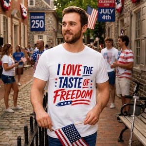Peut inclure: Un t-shirt blanc à manches courtes portant l'inscription I LOVE THE TASTE OF FREEDOM en lettres rouges et bleues, agrémenté de petites étoiles et d'un graphisme en coup de pinceau rouge. Porté avec un jean bleu et un petit drapeau américain.