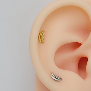 May include: Two small feather-shaped earrings, one gold and one silver, are shown in a close-up image of a model's ear.