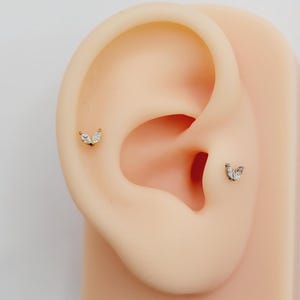 May include: Two gold ear piercings with small, clear, marquise-shaped stones. The piercings are in the cartilage of the ear.