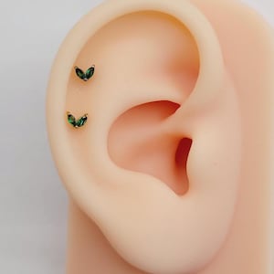 Puede incluir: Dos pequeños pendientes en forma de hoja verde con engaste dorado, mostrados en una oreja de maniquí.