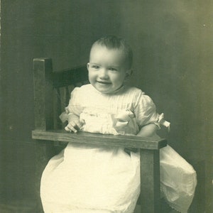 肘掛けに寄りかかって椅子に横向きに座っている幸せそうな赤ちゃん 1910 年代 RPPC 実写ポストカード写真