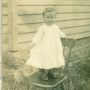 Menino de vestido parado do lado de fora na cadeira Fazenda Casa 1900 Retrato de estúdio Antigo RPPC Cartão postal com foto real