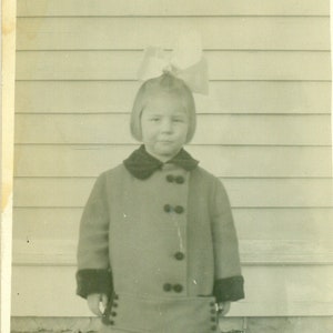 Op de afbeelding: Zwart-witfoto van een jong kind in een jas en rok. De jas is dichtgeknoopt en heeft een donkere kraag en manchetten. Een grote strik zit op het hoofd van het kind. Het kind staat op gras voor een gebouw.