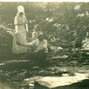 Lata 1910. Mężczyzna siedzący przy strumieniu i podający butelkę kobiecie w kapeluszu RPPC Prawdziwe zdjęcie pocztówki Czarno-białe zdjęcie