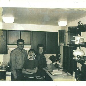 Puede incluir: Fotografía en blanco y negro de una familia en una cocina. La imagen muestra a un hombre, una mujer y un niño. La cocina tiene armarios oscuros, un fregadero y una encimera. Los miembros de la familia sonríen y miran a la cámara.
